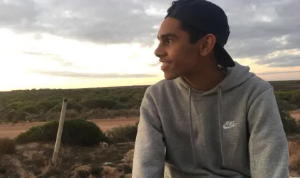 Young Aboriginal man wearing a grey hoody and cap looks out over bush landscape with cloudy sky in the background.