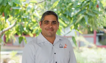 Indigenous man in light coloured shirt stands in front of green tree