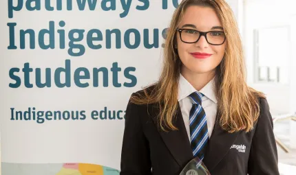 Young woman with light brown hair wearing glasses and black school jacket stands in front of a sign which says ‘pathways to Indigenous students, Indigenous education’.