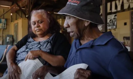 An Aboriginal woman in dress and dark cardigan sits next to an Aboriginal man in hat and blue shirt. In the background is a wall with posters and paintings on it.