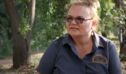 A head and shoulders shot of a woman with her hair pulled back, wearing glasses and a grey shirt. In the background are trees.