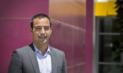 Man in grey suit standing in front of a maroon wall.