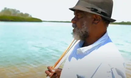 An Indigenous man in hat and pale shirt holds a spear over his shoulder. In the background is water and foliage.