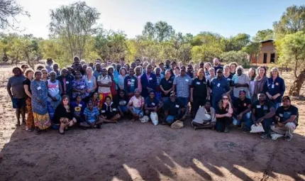 Large group of mainly Aboriginal people face the camera. They are dressed in casual clothing. Some stand and others sit on soft soil. In the background are trees and a building.