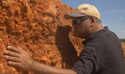 Aboriginal man in dark polo shirt, cap and sunglasses leans close to an ochre coloured rocky.