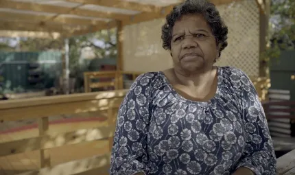 A mature woman in a patterned dress sits at a table. In the background is a fence and lattice and trees and more fence.