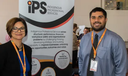 Indigenous woman in black jacket stands with Indigenous man in blue-grey shirt in a room with other people. Between them is a sign with logos and words including IPS Indigenous Professional Services.