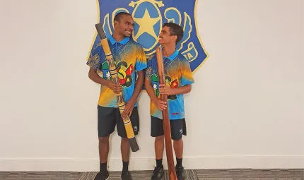 Two Aboriginal young men, wearing colourful shirts, hold didgeridoos as they stand beneath a blue and gold sign which includes a coat of arms.