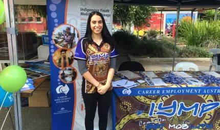 Young Indigenous woman standing in front of sign and to the side of a display table for Indigenous Youth Mobility Pathways. She is dressed in black pants and a colourful shirt showing Indigenous designs.