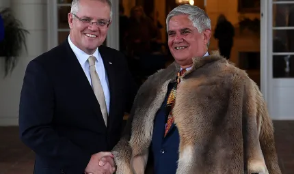 Non-Indigenous man in suit and tie at left and Indigenous man in ceremonial fur cloak and suit at right shake hands. In the background is an entrance to a building.