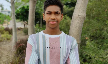 A young man in a colourful shirt with vertical stripes stands in front of some trees beyond which ae bushes and a road.