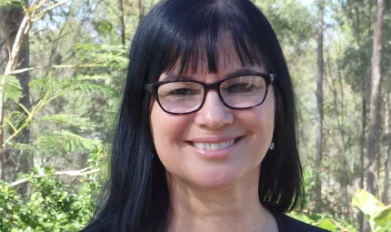 A woman with long black hair and wearing glasses faces the camera and smiles. In the background are trees and shrubs.