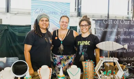 Three Aboriginal women stand behind a table covered in handmade products including necklaces and earrings. Behind them are posters with the words Gillawarra Arts on one and Gift of Our E on the other.