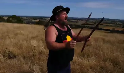 An Aboriginal man in black hat and black singlet with Aboriginal flag on it holds two sticks as he stands on a hill covered in dry grass. In the background is a blue sky, more hills and fields and trees.