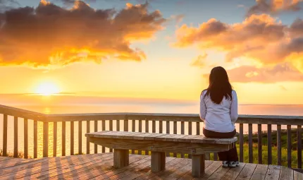 A woman with long dark hair and wearing a light jacket and dark pants sits on a bench overlooking water. In the background is a darkening sky, clouds and a setting sun.