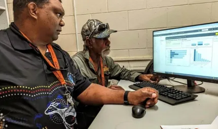 Two men dressed in casual clothing look at two graphs on a computer screen. In the foreground is a book and in the background is a brick wall.