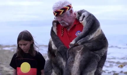 A young girl dressed in a t-shirt with Aboriginal flag design (black bar at top, red at bottom and yellow disc in middle) stands with an elderly man dressed in red shirt and fur cloak in front of a seascape.