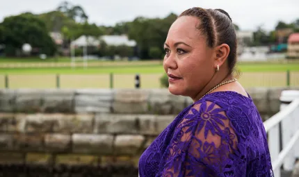 Indigenous woman with hair pulled back wearing a purple dress. In the background is a wall, then a playing field and buildings.