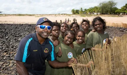 Aboriginal man in blue shirt and cap with Aboriginal youth in green shirts stand on rocks at a beach with sand and trees in the background. He supports an upright bamboo barrier.