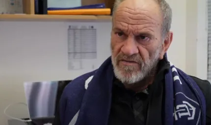 A middle aged balding man in dark jacket and purple scarf looks to the left. In the background is a shelf with various items on it and a document pinned to a white wall.