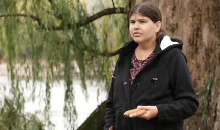 A woman in black top stands in front of a tree. In the background is a river and beyond that a riverbank and foliage.