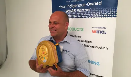 A man in a blue shirt holds a defibrillator as he stands in front of a poster which features the text: Integrity Health & Safety. Your Indigenous owned WH&S Partner.