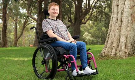 A young woman sits in a wheelchair on grass. In the background are trees.