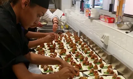 Indigenous woman dressed in black prepares multiple food dishes on stainless steel bench with fellow chefs in the background.