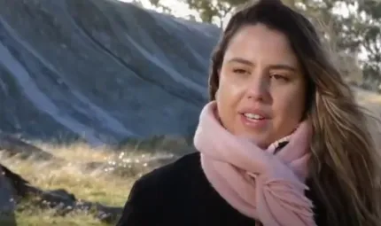 A head and shoulders view of a woman with long hair and wearing a black top and pink scarf. In the background is grass, a large long rock and trees.