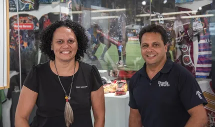 An Indigenous woman dressed in black and an Indigenous man in a dark shirt look to camera. In the background is a display of shirts, boots and posters.