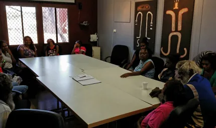 A group of Indigenous people sitting round a table in a meeting room with Indigenous art on the walls and window at one end.