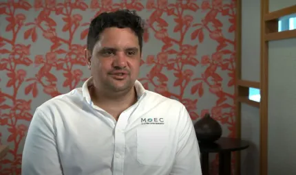 A young adult man with dark hair and wearing a white shirt sits in a room with an ochre red design on a pale wall. To his left and behind him is a small table with a sculpture on it.