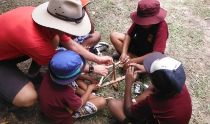 Man in hat teaches four Indigenous students in hats and maroon shirts how to start a fire