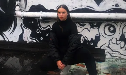 A young woman with long brown hair dressed in dark top and pants, and white shoes squats in front of a wall painted with black and white images. A pipe runs horizontally across the wall.