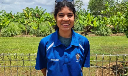 Young woman in blue shirt stands in front of a metal fence. In the background is grass and trees and a cloudy sky