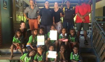 A group of Aboriginal school students sit on steps, holding a letter from the Prime Minister the Hon. Malcolm Turnbull, MP. Behind them stand five adults.