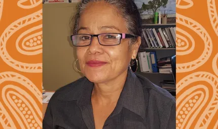 Indigenous woman with hair drawn back and wearing glasses and a dark shirt. Behind her is shelving and other office content.