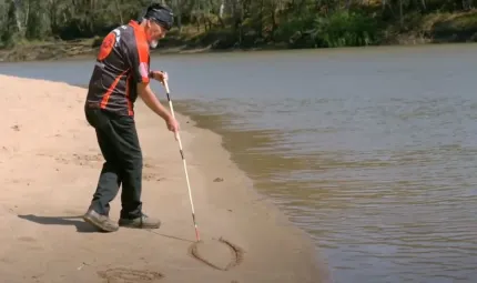 A middle-aged man in dark trousers and red and black shirt stands next to a river and holds a long stick with which he draws lines in the soft sand bank. In the background is more of the river and trees on the opposite bank.