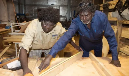 Two Indigenous men working at a wood working bench with the elder man directing the younger