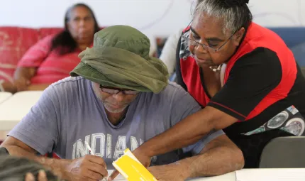 Indigenous man with green hat and grey t-shirt writes on a paper as an Indigenous woman in red and black apparel points at the paper. Another Indigenous woman sits on a couch in the background.