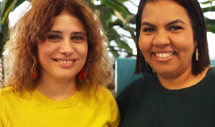 Two smiling women stand shoulder to shoulder. The woman at left has light brown hair and wears a yellow t-shirt. The woman at right has dark brown hair and wears a dark t-shirt. In the background is a tree.