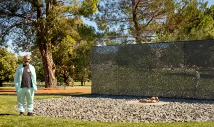 A man in pale blue suit and dark shirt stands outside on a sunny day in front of circle of rocks and reflecting wall. In the background are trees and lawns.