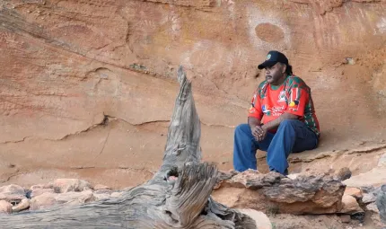 Aboriginal man in red shirt, blue jeans and a black cap sits on a rock ledge behind which are cave paintings. In front of him is a fallen log.