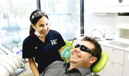 Young Indigenous woman in dark blue uniform stands holding a dentist drill behind a smiling man wearing sunglasses and lying back in a dentist chair.