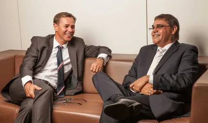 Man on left in brown suit and another man on right in dark grey suit sit on a brown couch with legs crossed and smiling.