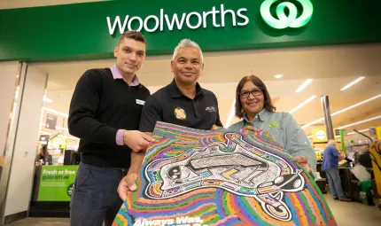 Three people hold a poster featuring an image of the Australian landmass, in front a Woolworths store. Above is the Woolworths name and logo. In the background is the store with shoppers.