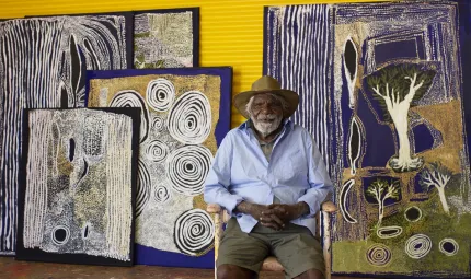 An elderly man in blue shirt, khaki shorts and a broad brimmed hat sits on a chair in front of a group of paintings leaning up against a yellow wall. The paintings feature swirls and lines.