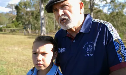 Elderly Aboriginal man in blue polo shirt with Aboriginal designs on the sleeves and wearing a cap stands with a young girls in a blue shirt. In the background is grass and trees.