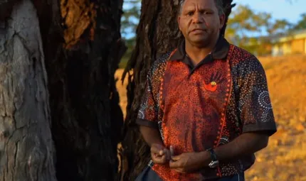 An Aboriginal man with short hair and wearing a black and orange polo necked shirt looks at camera.