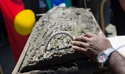 The hand of an Aboriginal man, wearing a silver watch, gently lays on top of a traditional wood box. The Aboriginal and Torres Strait Island flags can be seen in the background.